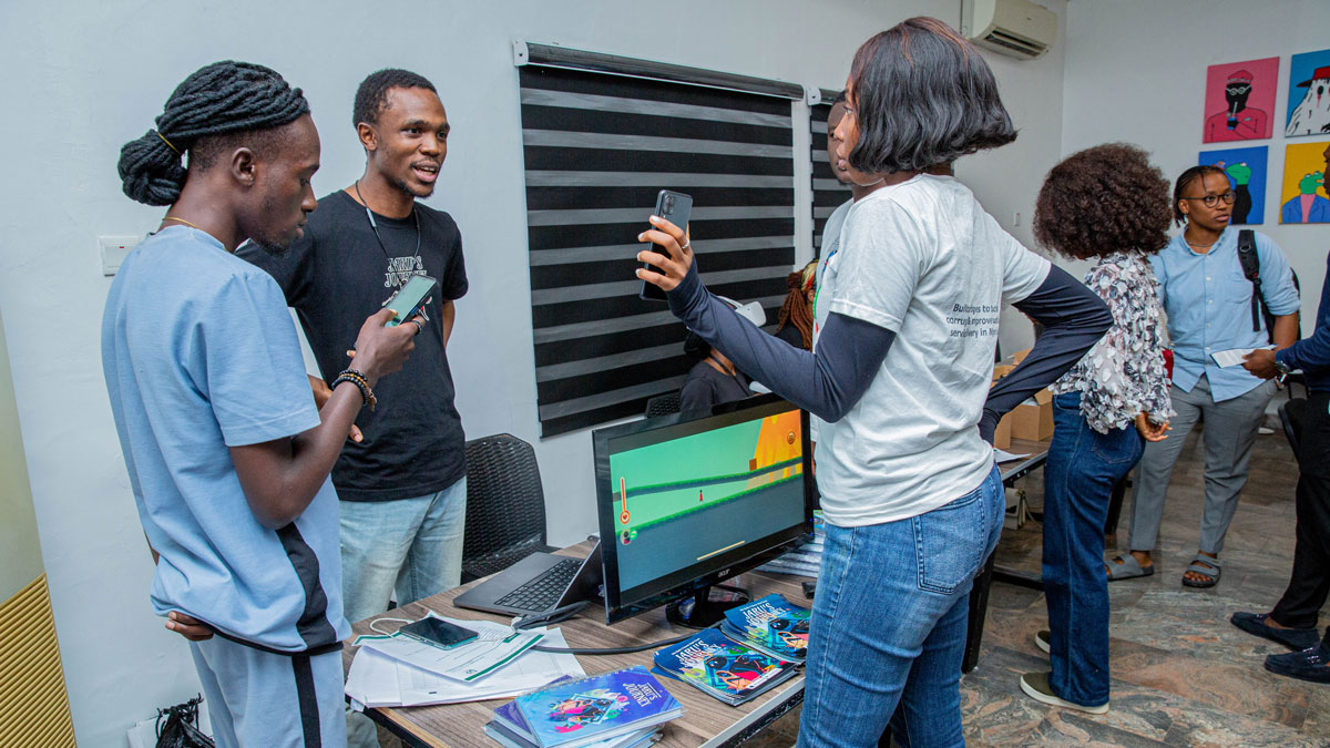 People interacting around a table with a computer, books, and a computer monitor.