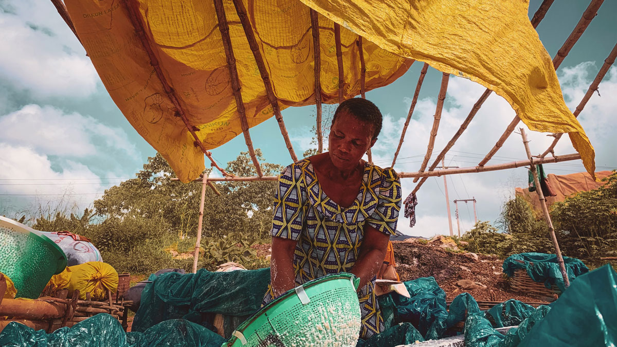 A person under a yellow fabric shelter, wearing a patterned dress and working with a green basket amidst greenery.