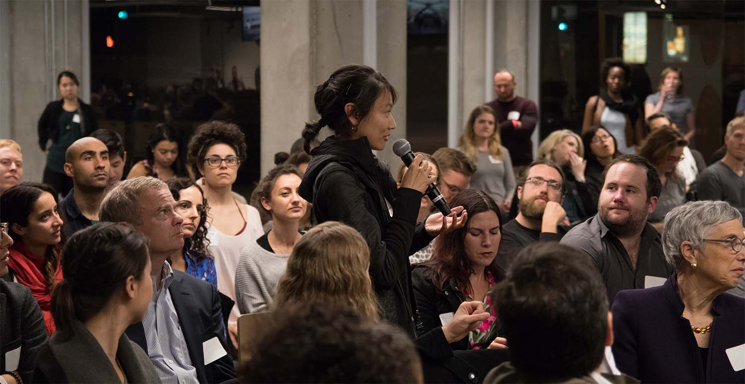 A woman stands and speaks into a microphone in a crowded room of seated and attentive people.